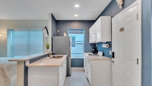 A compact kitchen with white cabinets, a built-in oven, and a window with blinds. - The Coral House Motel (Hollywood)