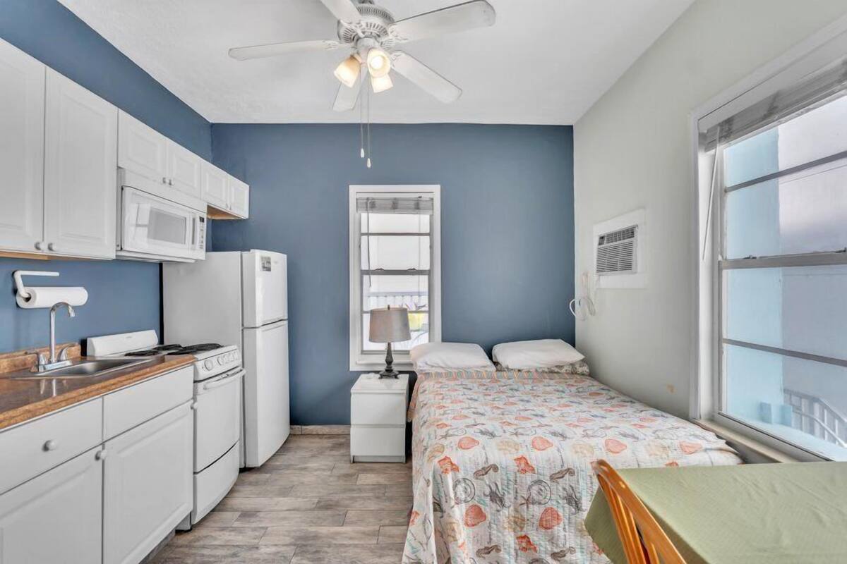 A compact room with a kitchenette, a bed with floral bedding, a ceiling fan, and a window with blinds.