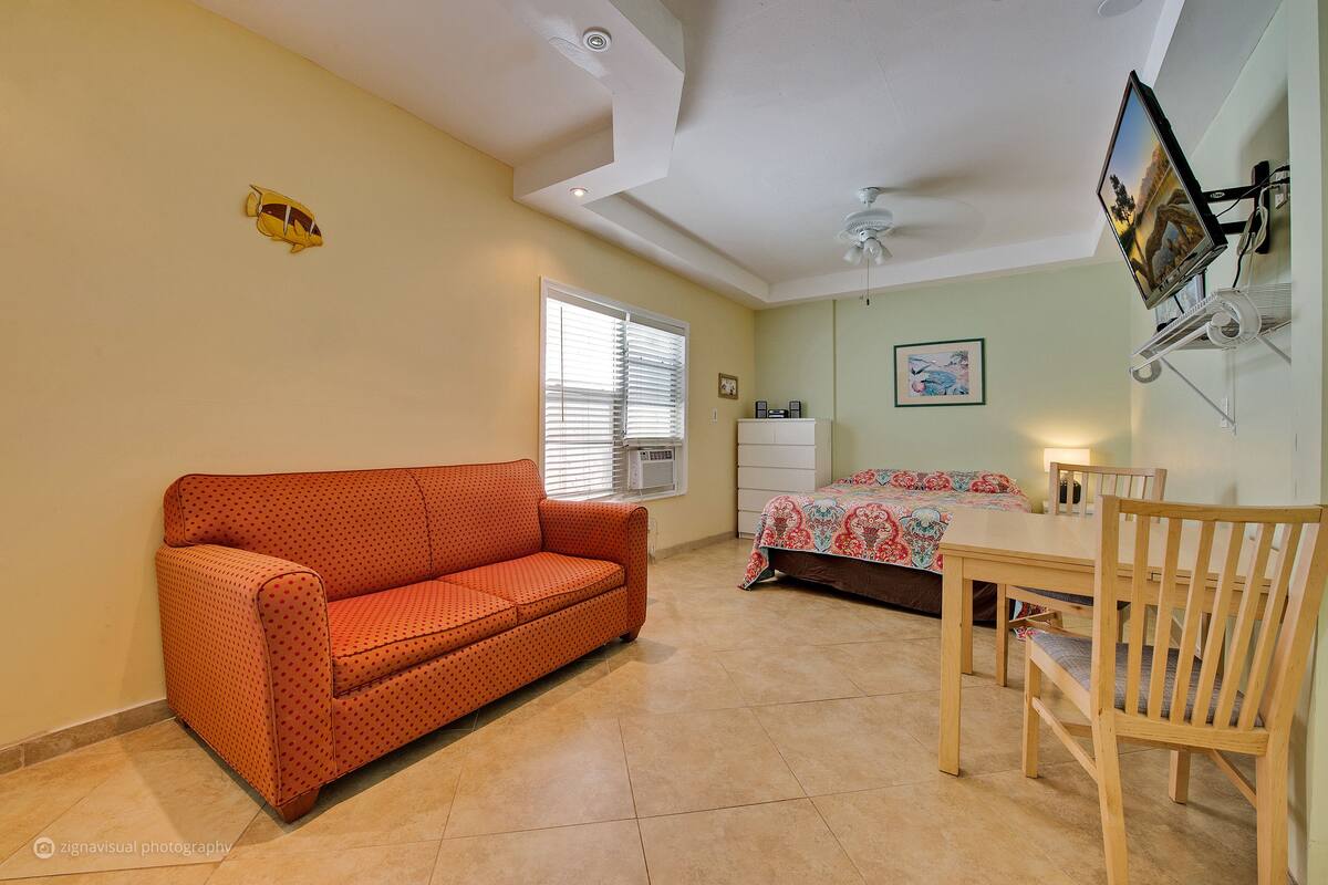 A room with a sofa, a dining table with chairs, a ceiling fan, and a wall-mounted TV.
