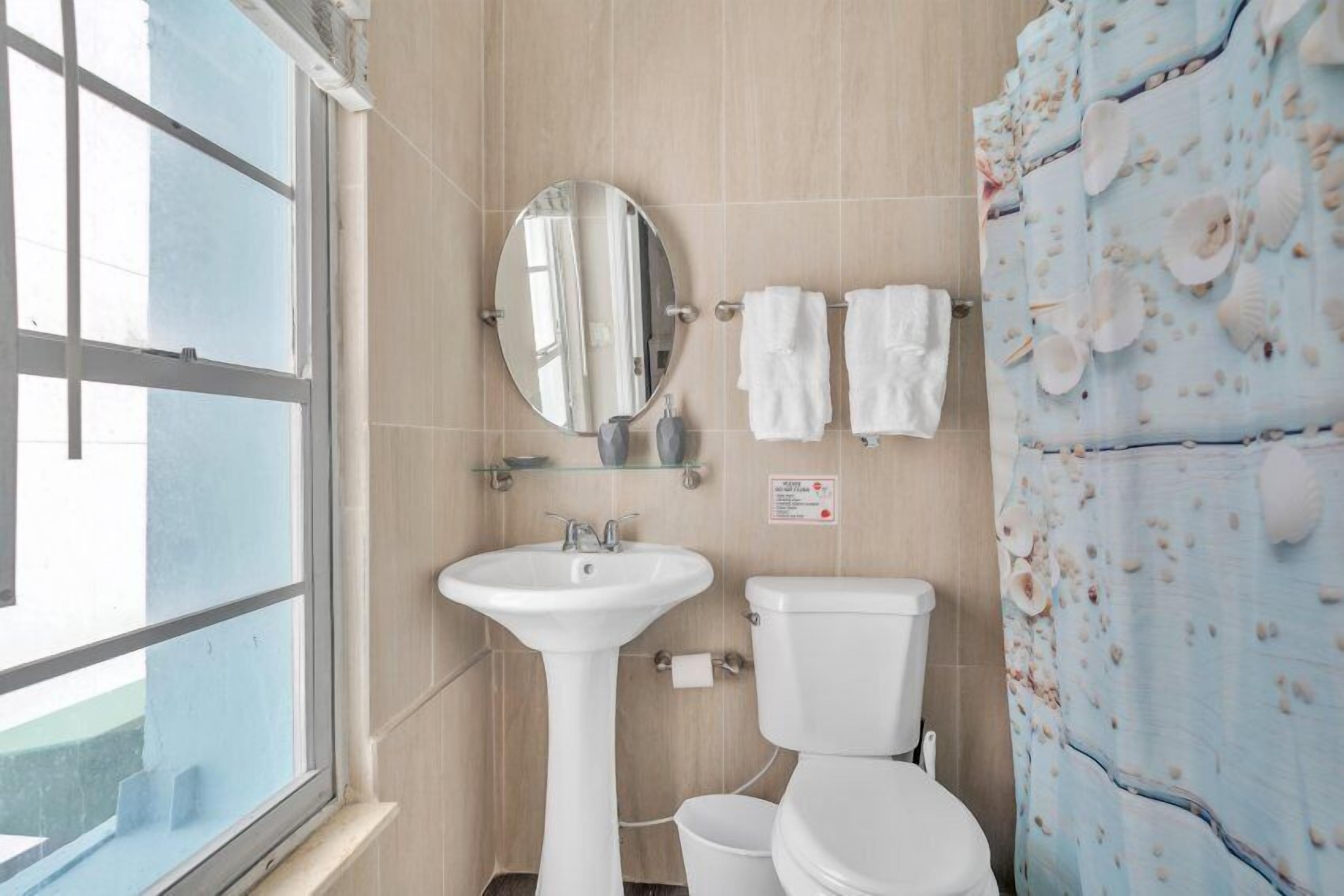 Un baño con ventana, lavamanos, inodoro y cortina de ducha con estampado de conchas.