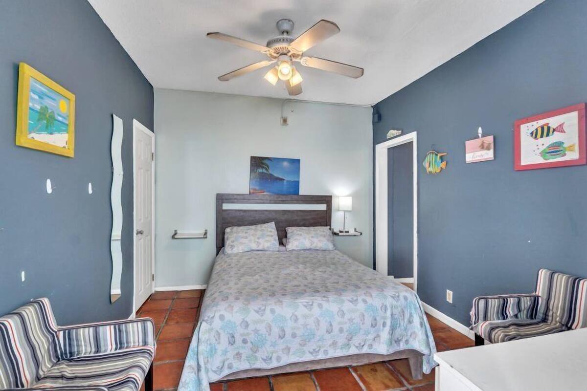 A bedroom with a bed, two chairs, a ceiling fan, and wall art.