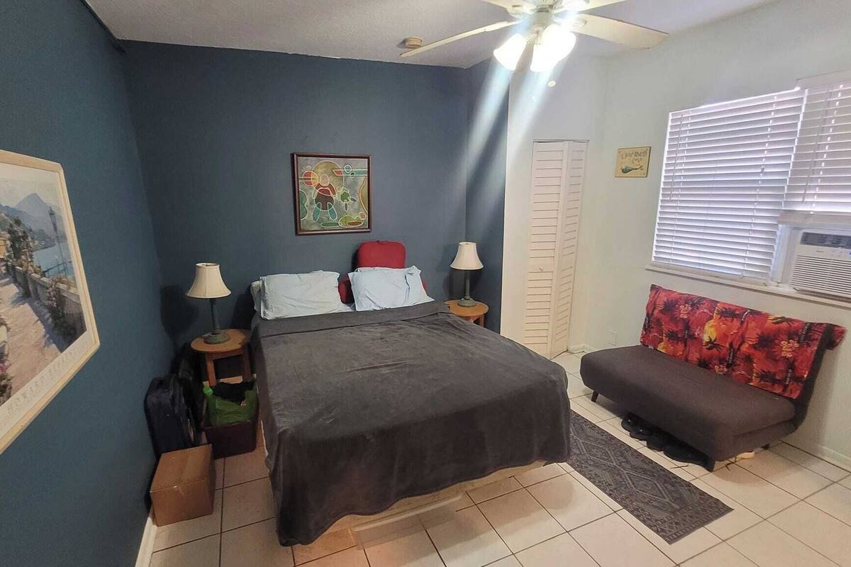 A bedroom with a bed, a sofa, a ceiling fan, a window with blinds, and a painting on the wall.