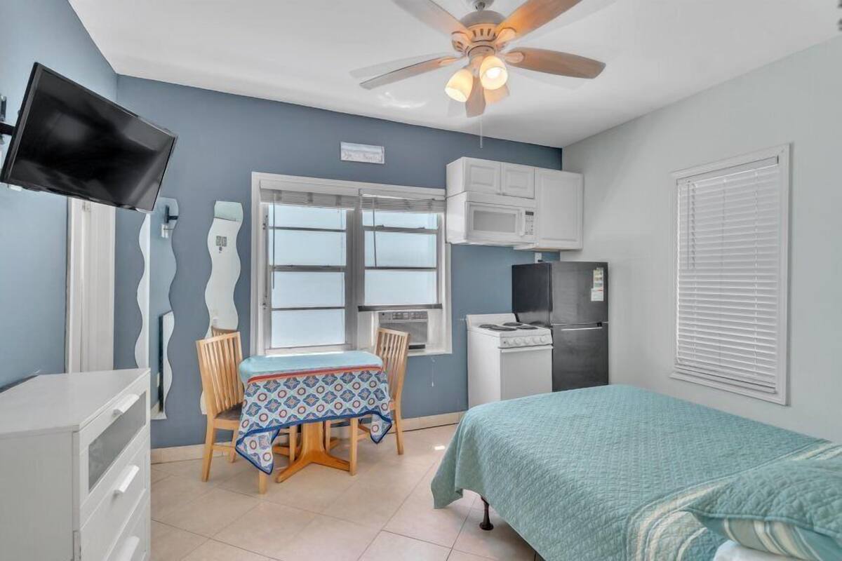 A compact room with a bed, a dining table with chairs, a kitchenette, a ceiling fan, and a TV mounted on the wall.