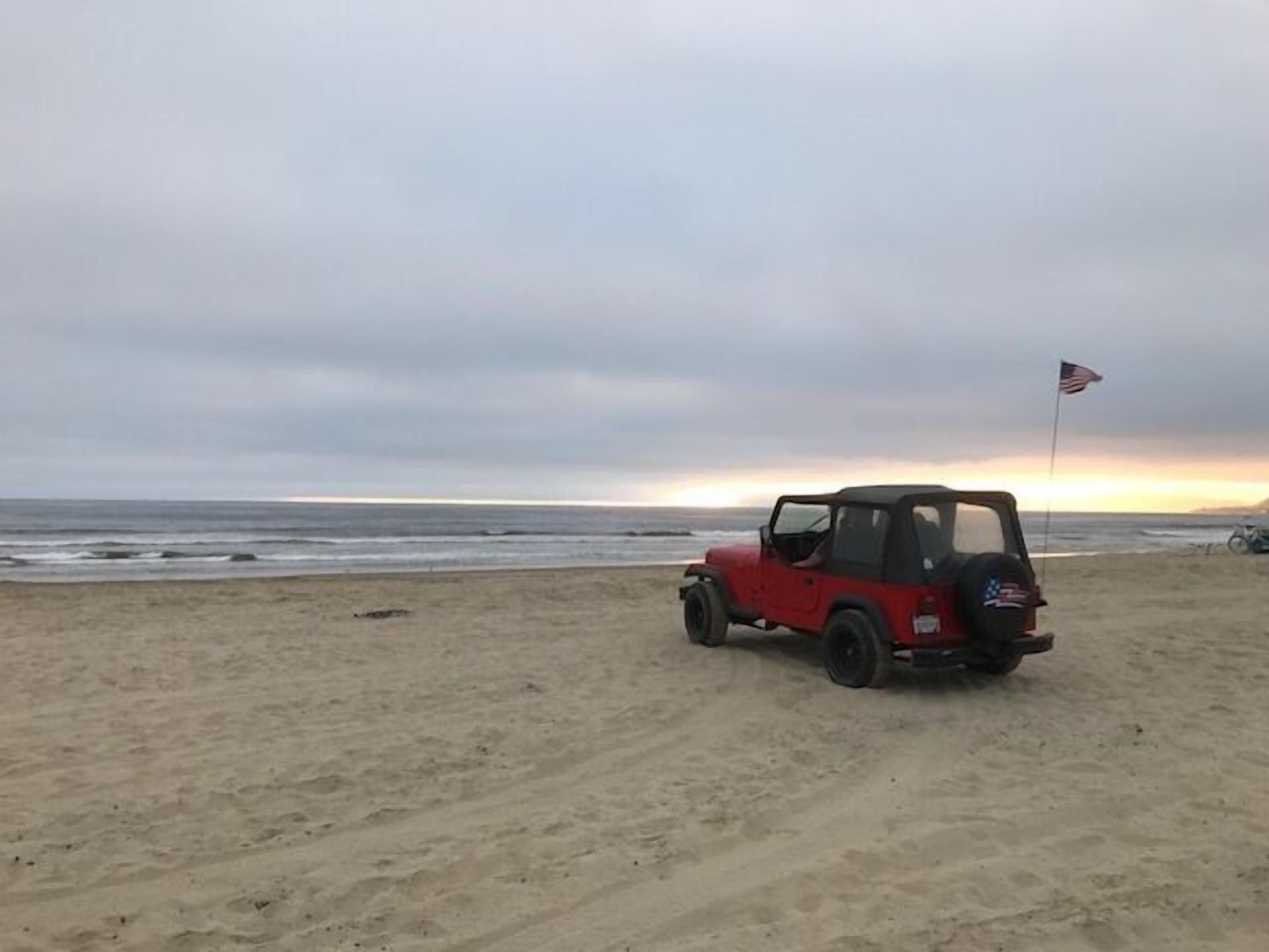 A red off-road vehicle parked on a sandy beach with an American flag on the back.