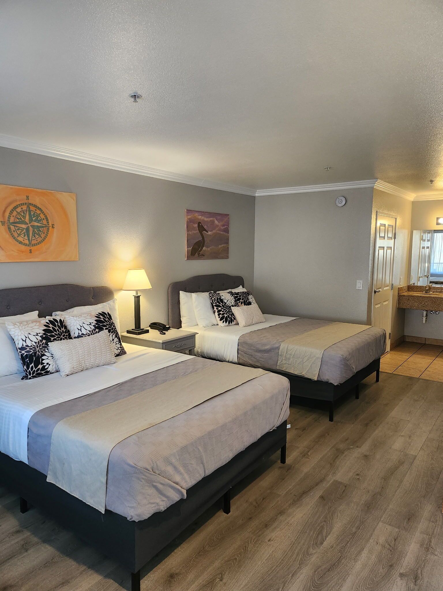 A hotel room with two beds, a bedside table with a lamp, a wall clock, and a painting.