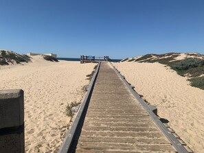 Una pasarela de madera que conduce a una playa con dunas y un cielo azul despejado.