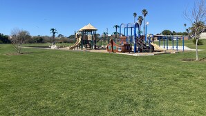 A playground with slides and climbing structures, surrounded by a grassy field and palm trees.