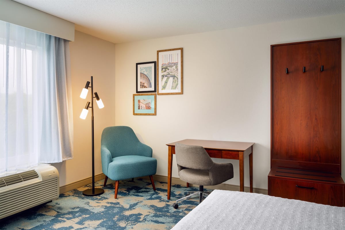 A hotel room with a bed, two chairs, a desk, a wardrobe, a floor lamp, and framed pictures on the wall.