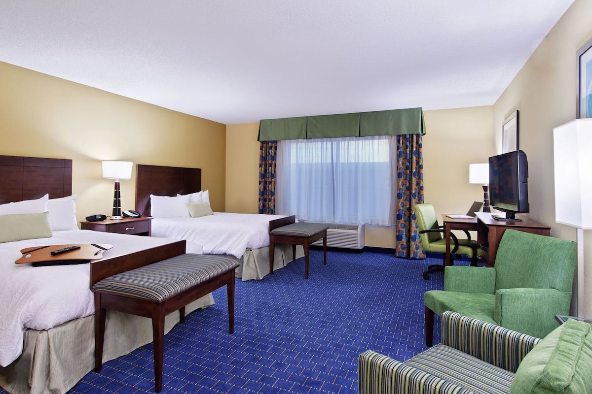 A hotel room with two beds, a desk with a computer, a TV, and a seating area with chairs.
