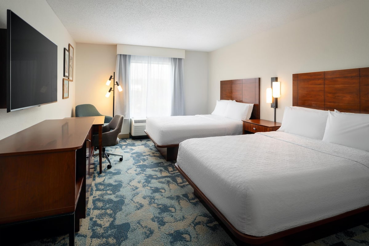 A hotel room with two beds, a desk, a chair, a flat-screen TV, and a large window with curtains.
