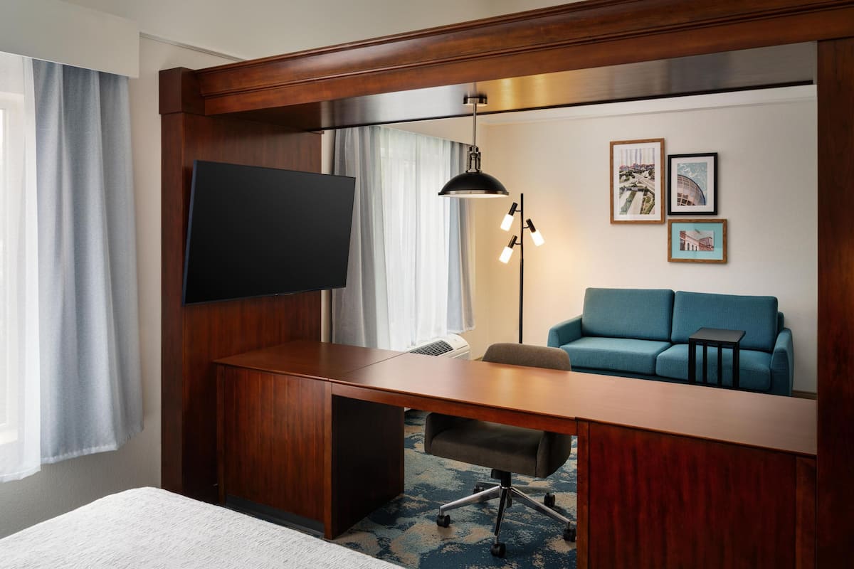A hotel room with a wooden desk, a flat-screen TV, a sofa, and a standing lamp.