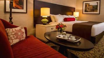 Habitación de hotel con cama, mesita de noche, una mesa de centro con tazas y flores, y un sofá rojo.
