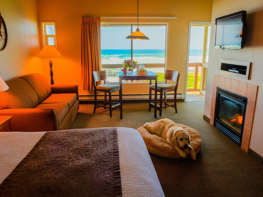A hotel room with a bed, a sofa, a dining table with chairs, a fireplace, a TV, and a view of the ocean.