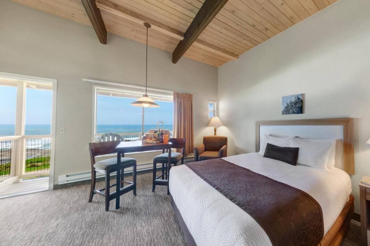 A hotel room with a large bed, a desk with two chairs, a view of the ocean, and a wooden ceiling.