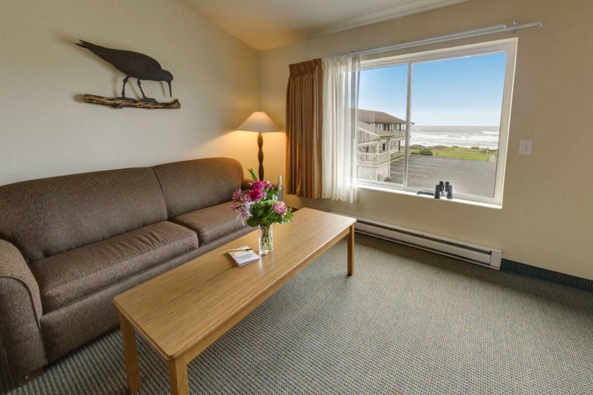A hotel room with a brown sofa, a wooden coffee table with a vase of flowers, a lamp, and a large window with a view of a building and the ocean.