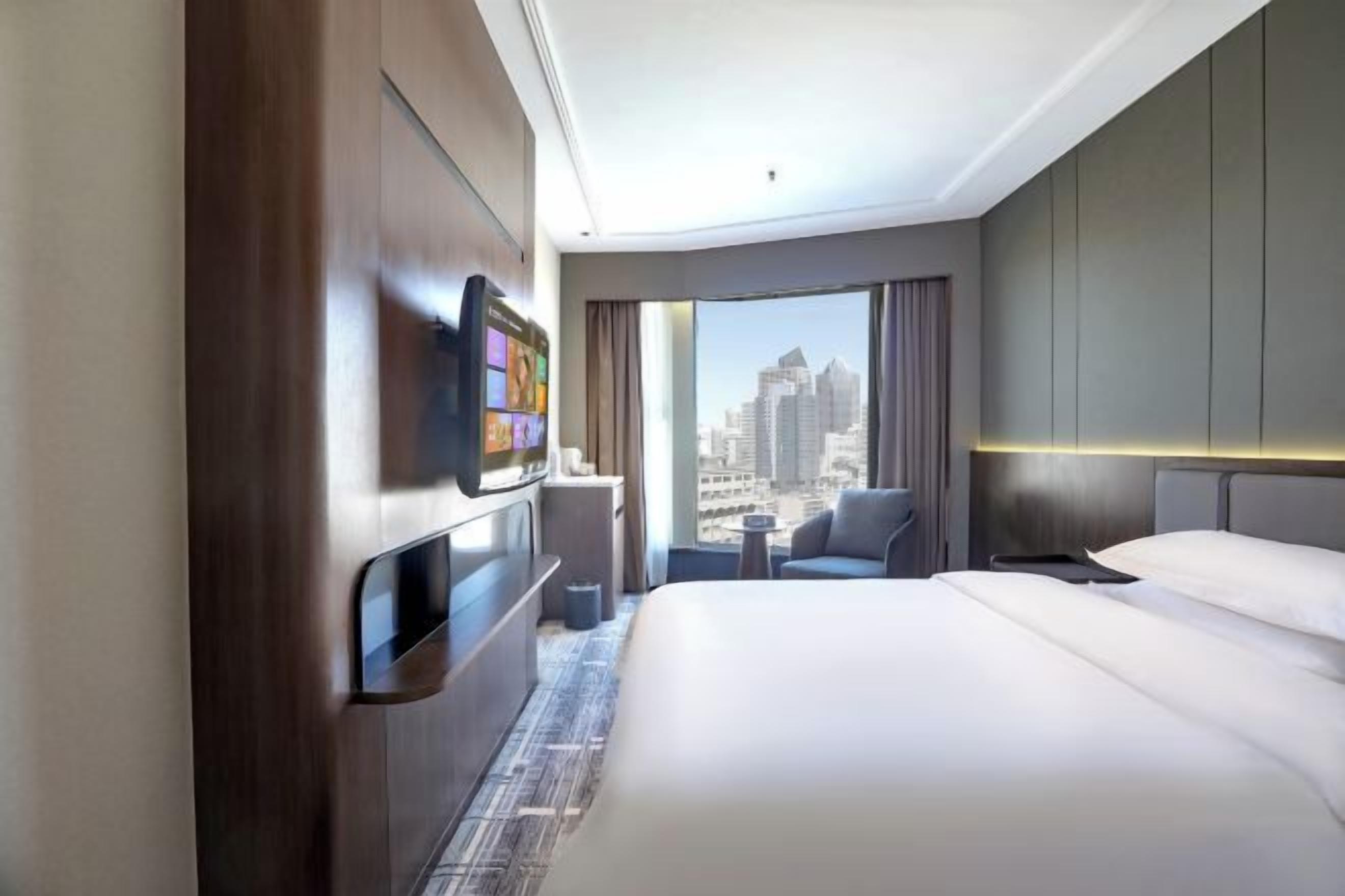 A hotel room with a bed, a TV mounted on the wall, a small table, a chair, and a view of the cityscape through large windows.