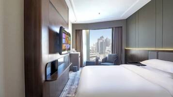 A hotel room with a bed, a TV mounted on the wall, a small table, a chair, and a view of the cityscape through large windows.