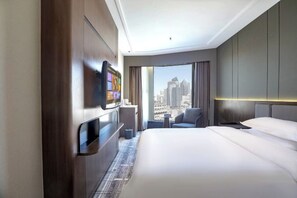 Habitación de hotel con cama, televisión montada en la pared, una mesita, una silla y vistas a la ciudad a través de amplias ventanas.