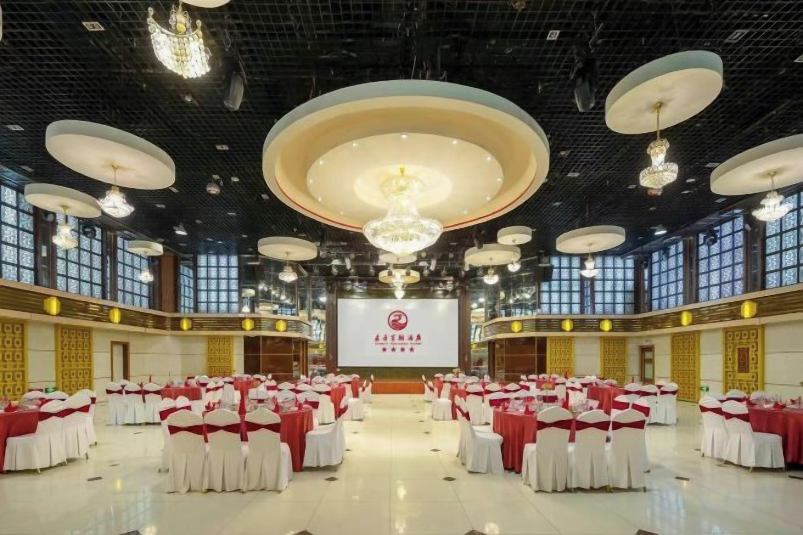 A large hall with rows of tables and chairs, a stage with a screen, and chandeliers.