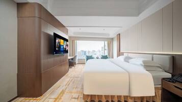 A hotel room with a large bed, a TV mounted on the wall, a desk with a lamp, and a view of the city through the window.