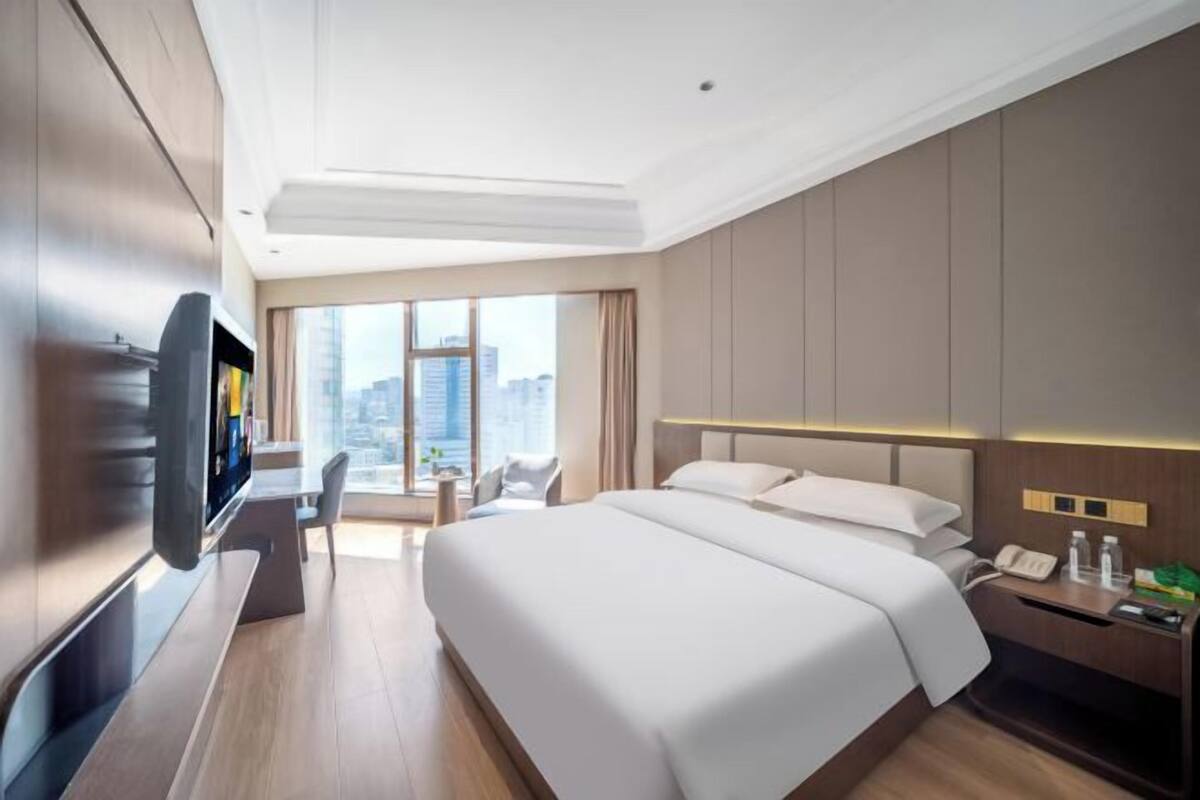 A hotel room with a large bed, a desk with a flat-screen TV, a bedside table with a phone and toiletries, and a view of the city through large windows.