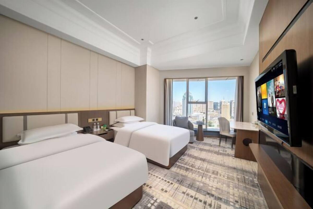 A hotel room with two beds, a desk, a TV, and a large window with a city view.