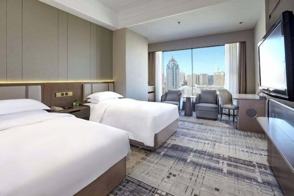 A hotel room with two beds, a desk, a chair, a TV, and a large window offering a city view.