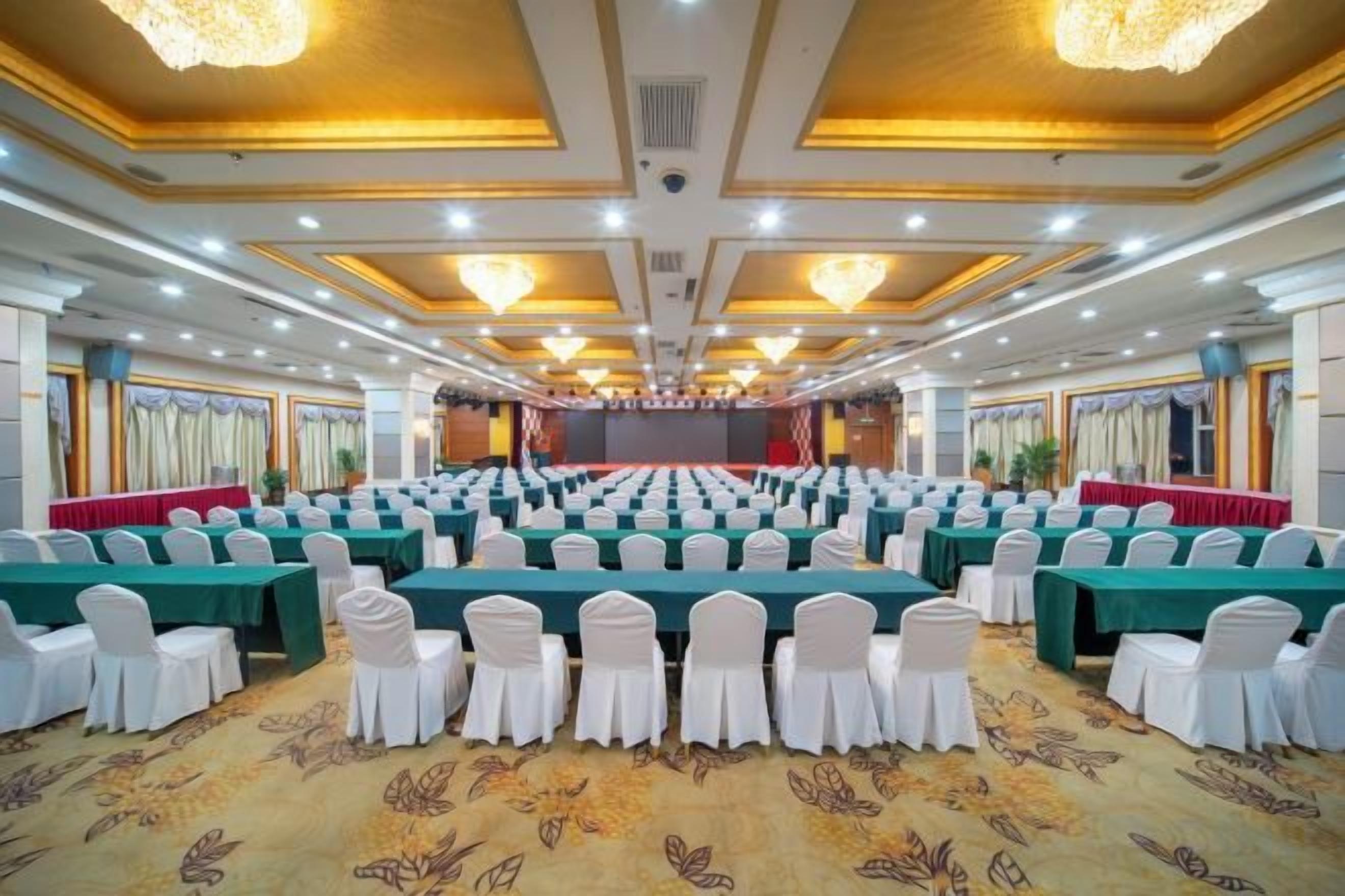 A conference room with rows of tables and chairs, a stage at the front, and a carpeted floor.