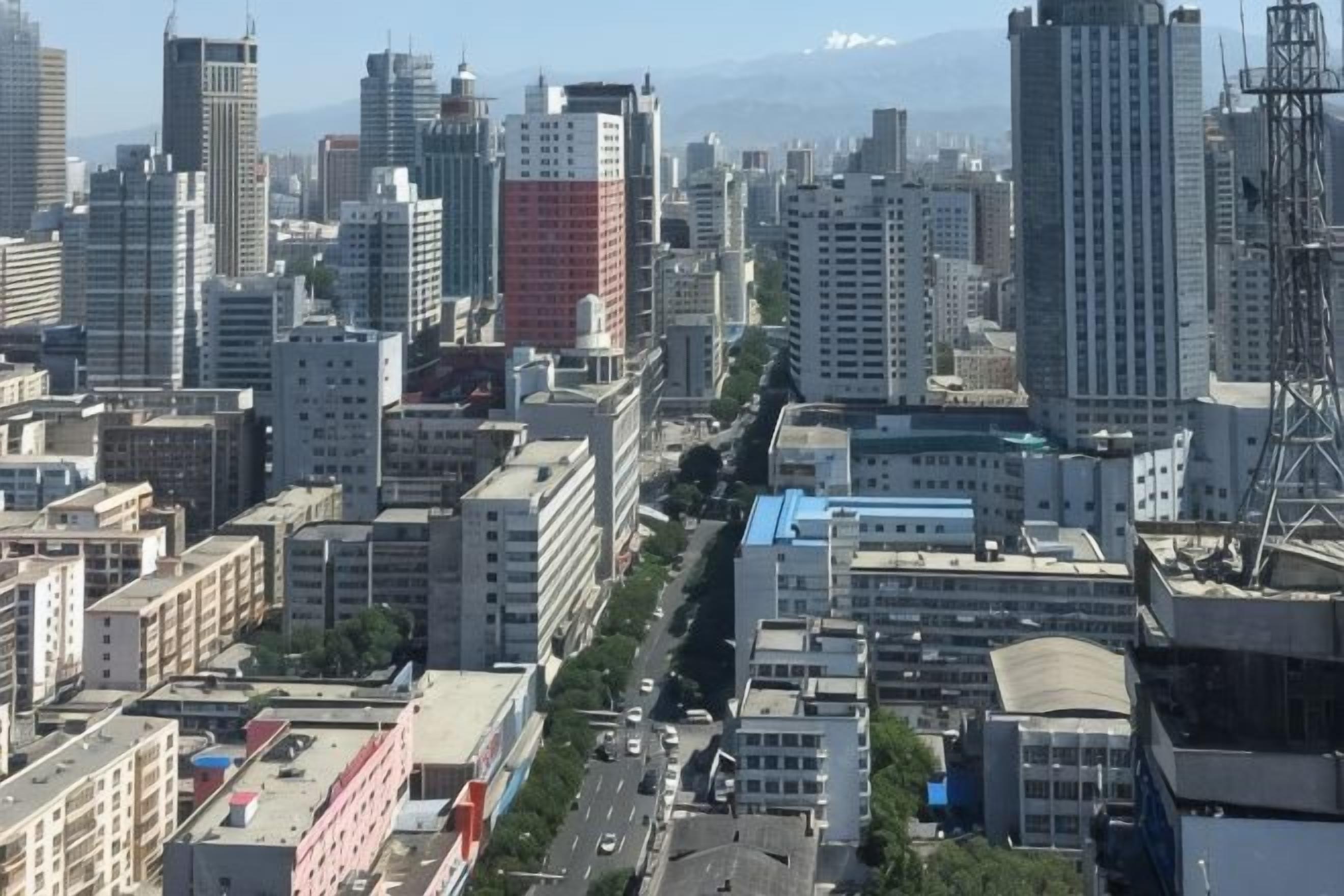 Un paisaje urbano con edificios altos, cielo despejado y una cadena montañosa en la distancia.