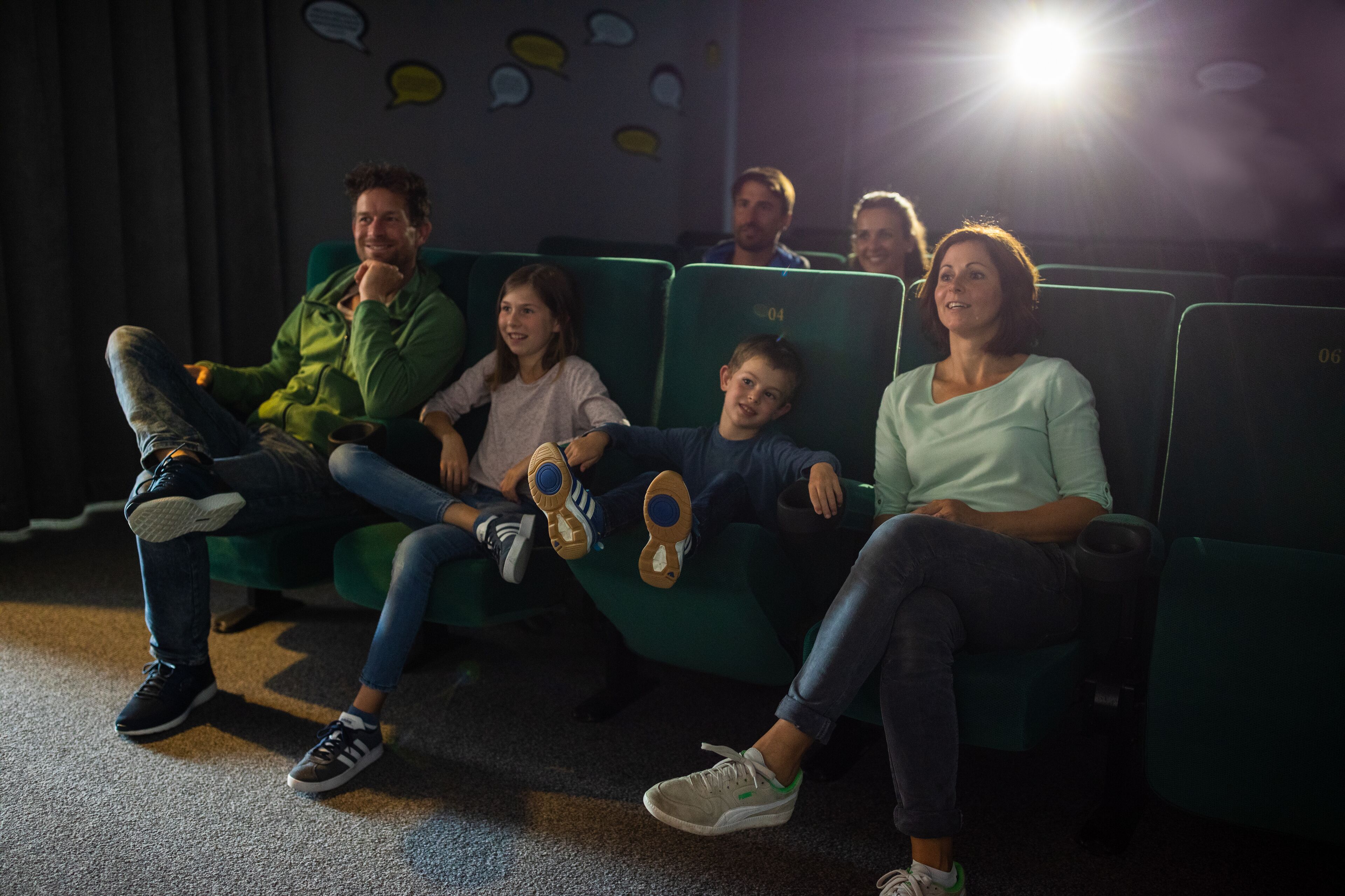 A group of people seated on green chairs in a dimly lit room with a bright light source in the background.
