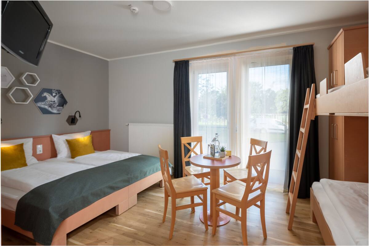 A hotel room with a bed, a small table with chairs, a TV mounted on the wall, and a window with curtains.
