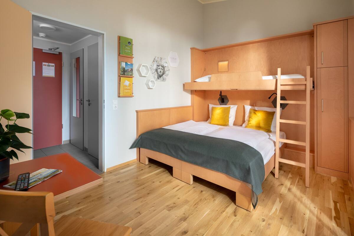 A bedroom with a bunk bed, wooden flooring, a desk with a chair, and a potted plant.