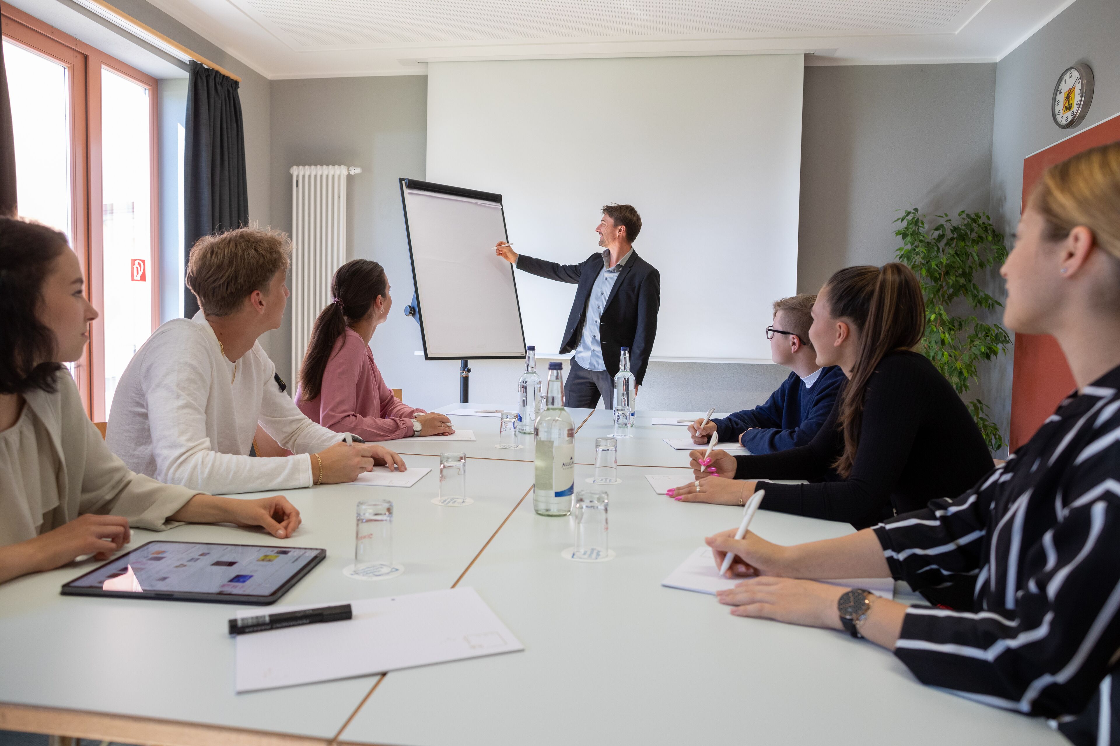 A conference room with a whiteboard, a presenter, and attendees seated around a table with water bottles and notepads.