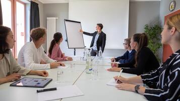A conference room with a whiteboard, a presenter, and attendees seated around a table with water bottles and notepads.