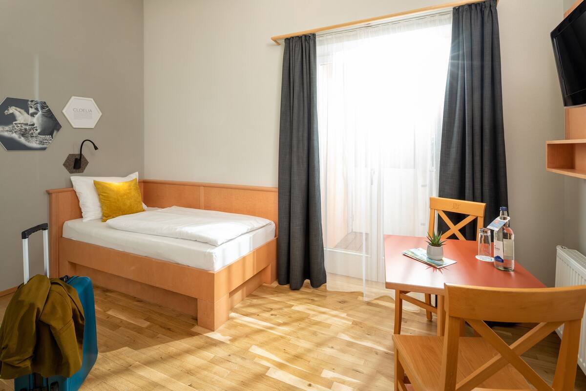 A hotel room with a bed, a table, a chair, a suitcase, and a TV on a stand.