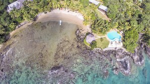 Vue aérienne d’un complexe hôtelier tropical doté d’une plage, d’une piscine et de plusieurs hébergements de style bungalow, le tout entouré d’une végétation luxuriante.