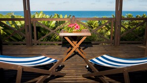 Une terrasse en bois avec deux chaises longues à rayures et une petite table agrémentée de fleurs, offrant une vue sur un paysage tropical et l’océan.