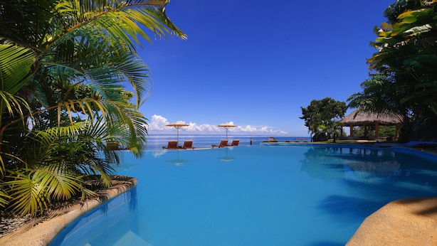 Une magnifique piscine à débordement d’un bleu profond, agrémentée de chaises longues et de parasols, entourée d’une végétation tropicale luxuriante et d’une structure au toit de chaume.
