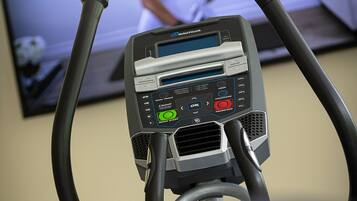 Un vélo d’appartement doté d’un écran numérique et d’un panneau de commande, un téléviseur en arrière-plan diffusant une émission sur une personne qui fait de l’exercice, et un appareil fixé au mur avec une interface circulaire.