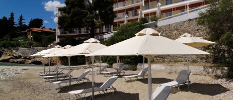 A beach with sun loungers and umbrellas, a stone building in the background, and a clear blue sky.