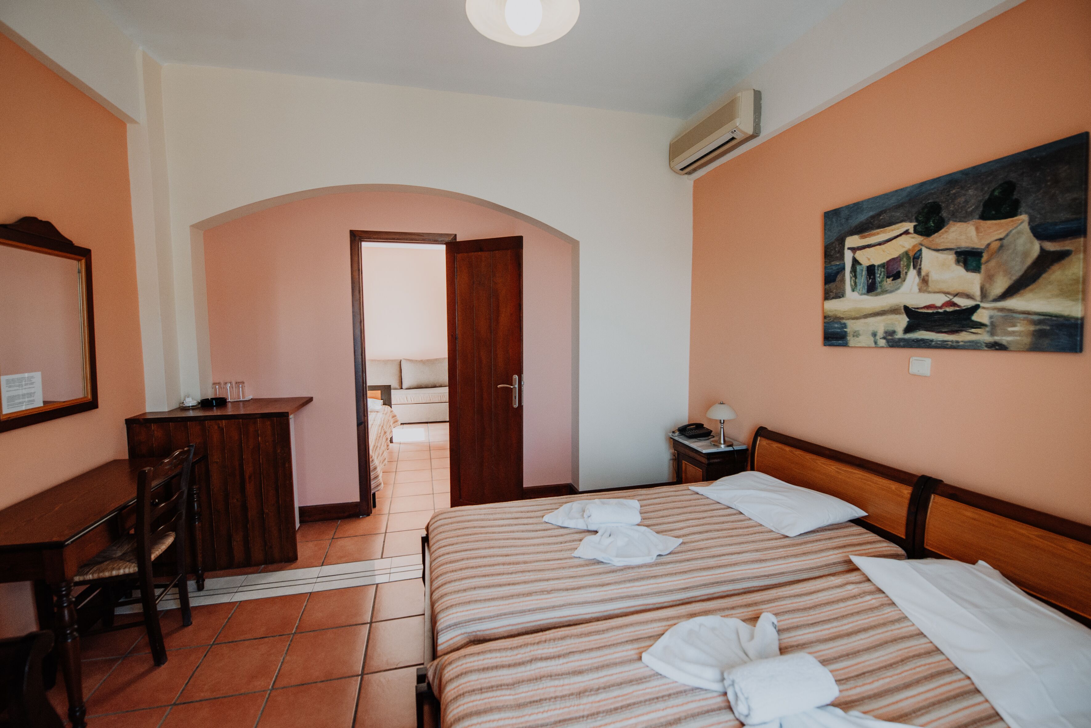 A hotel room with a bed, a desk, a chair, a painting on the wall, and an arched doorway leading to another room.