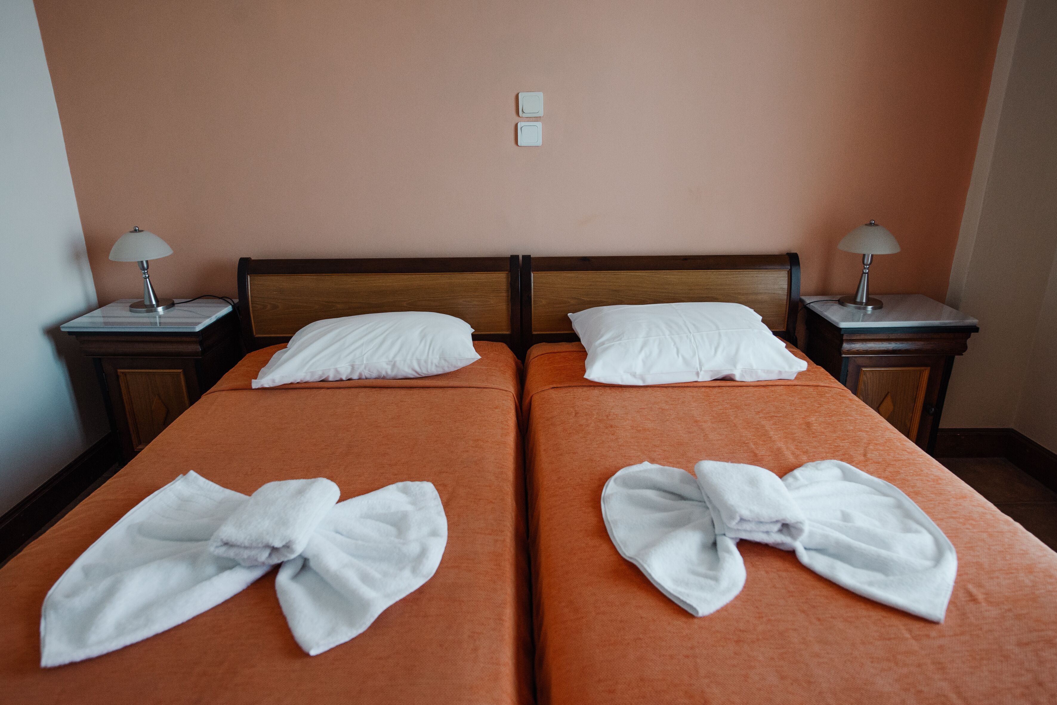 A hotel room with two beds, each with a white towel folded on the bed. There are two bedside tables with lamps, and the walls are painted peach.