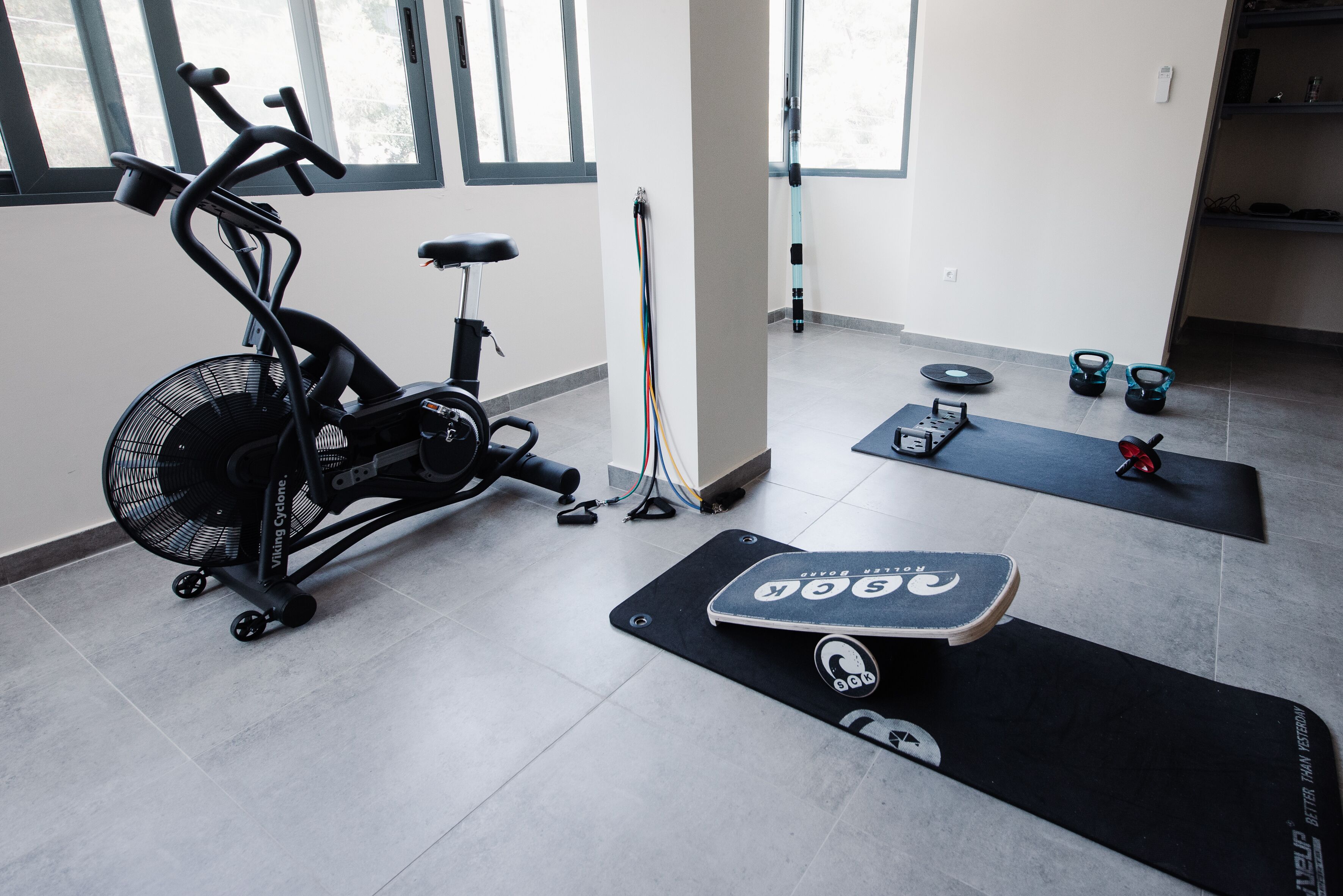 Ein Heimfitnessraum mit einem Ergometer, einem Ventilator, einer Yogamatte und Kettlebells.