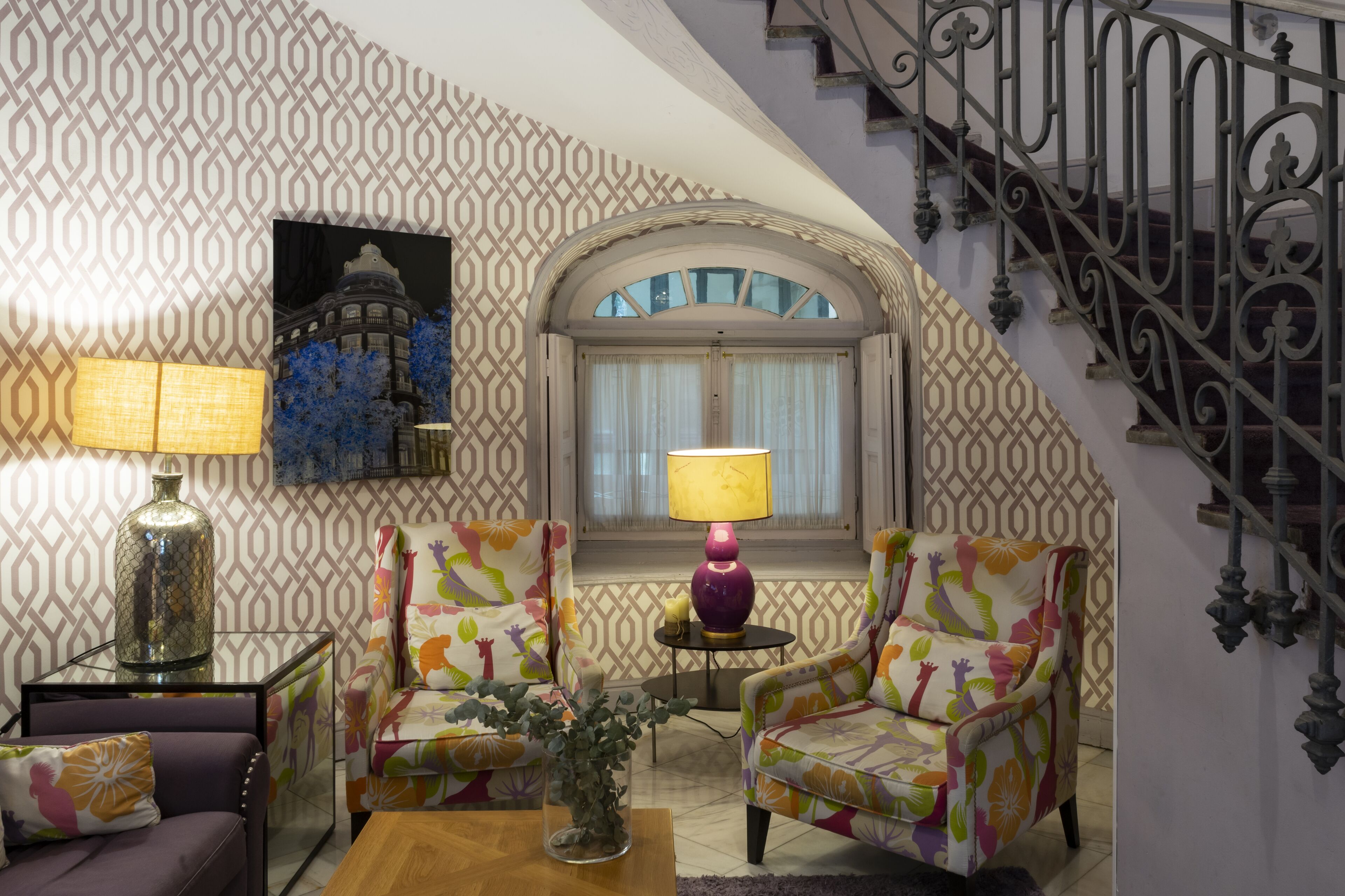 A living room with patterned wallpaper, two colorful armchairs, a table with a lamp, and a staircase with ornate railings.