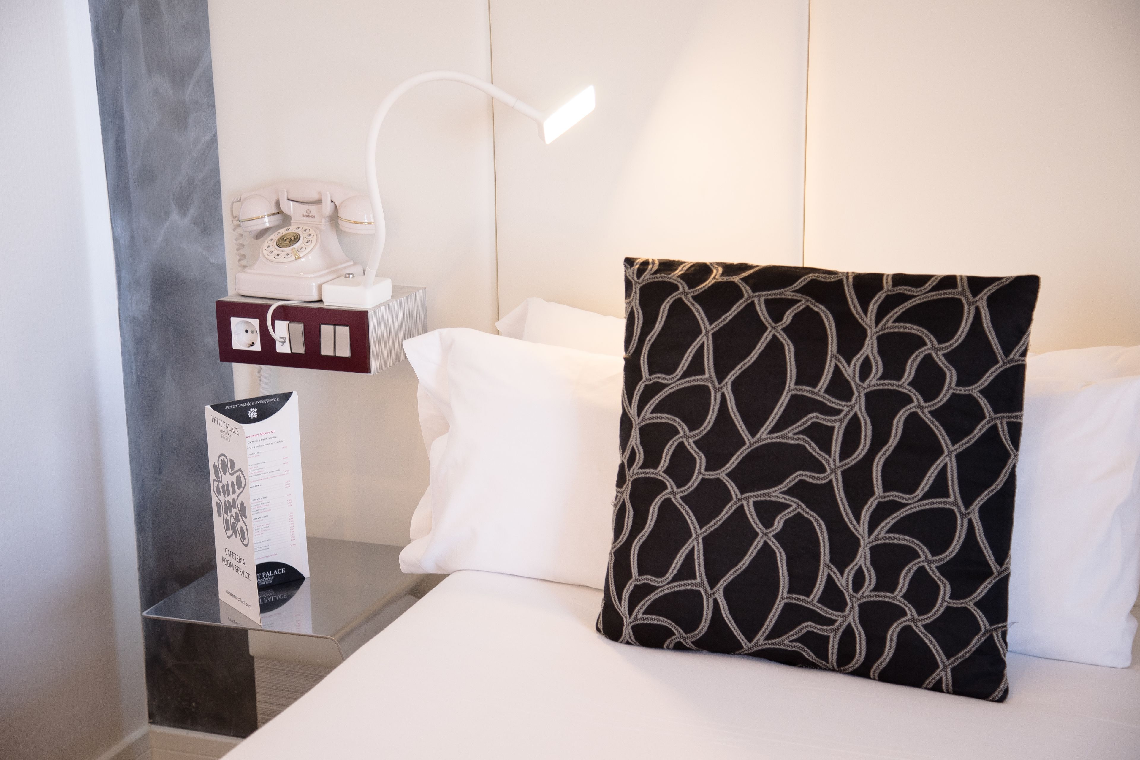 Una habitación de hotel con una cama, almohadas blancas, una almohada negra estampada, una mesita de noche con un teléfono y un folleto, y una lámpara moderna.