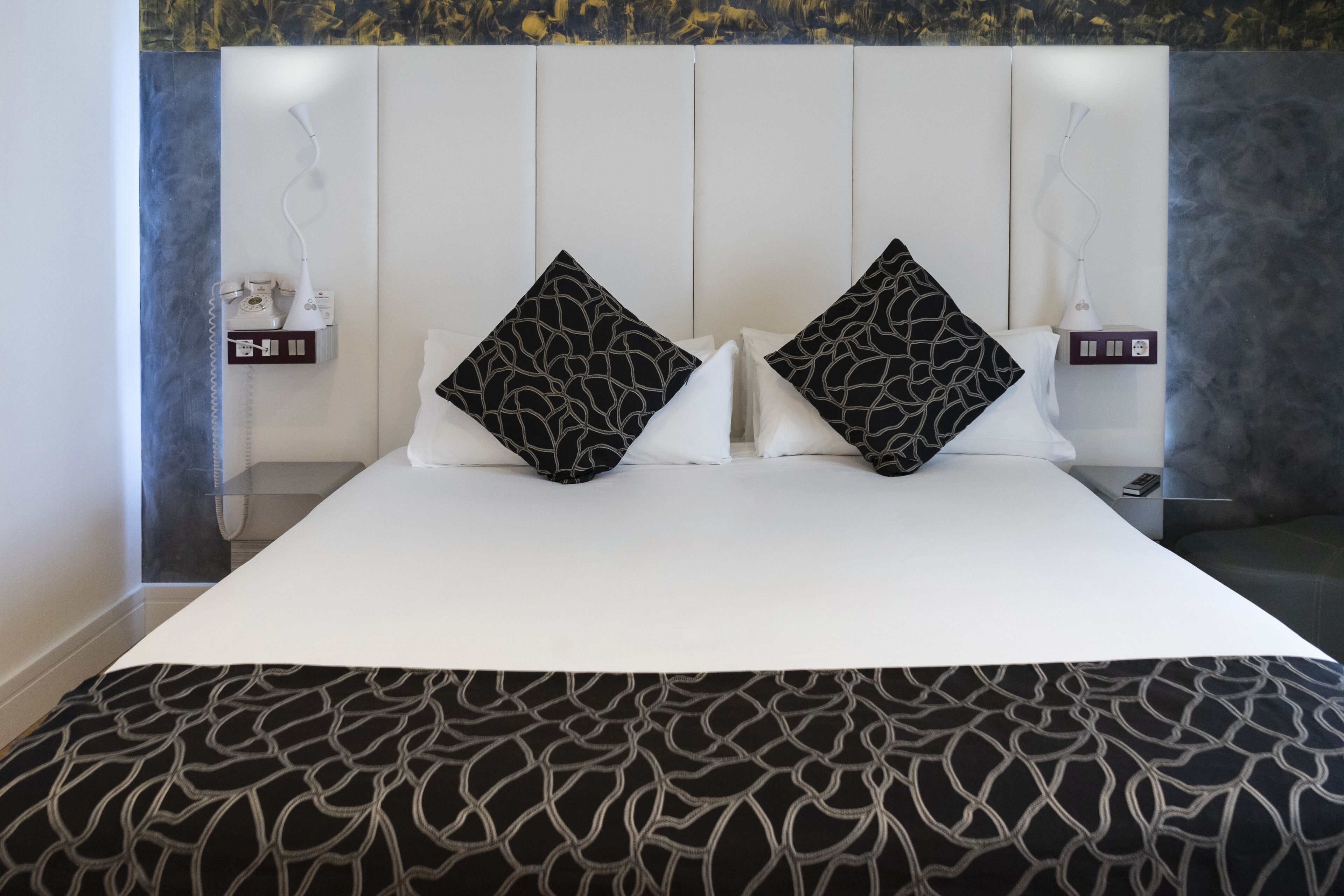 Ett modernt hotellrum med en stor säng, två mönstrade kuddar, en vit sänggavel och ett mönstrat överkast.