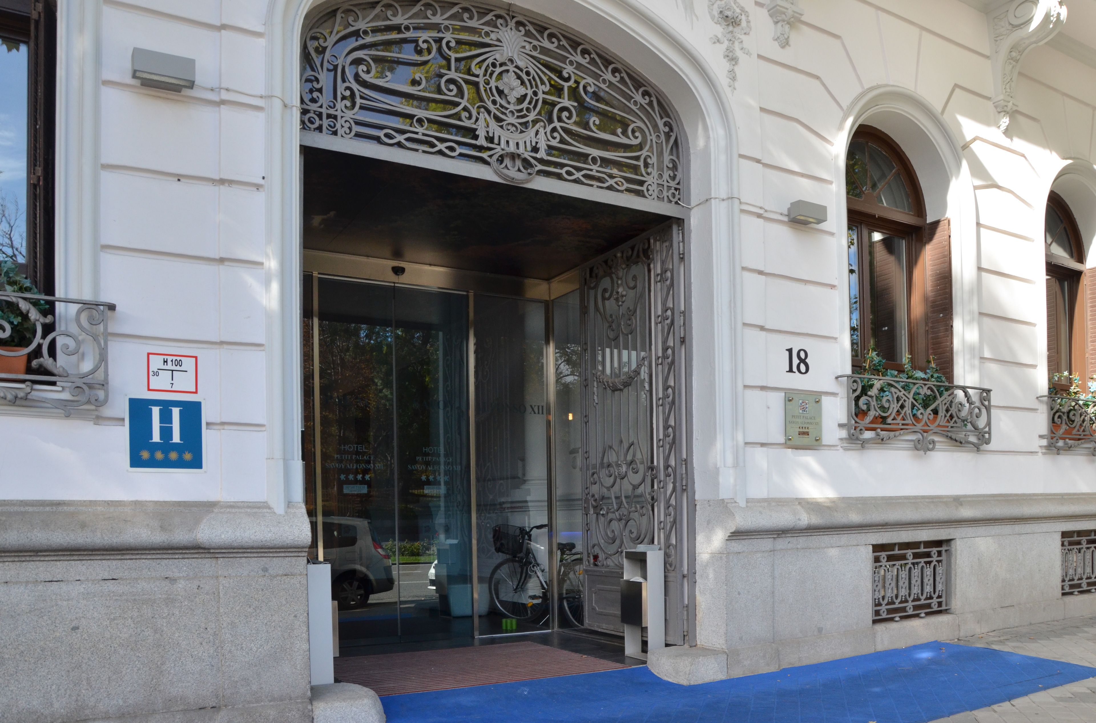 L’entrée d’un hôtel, ornée de ferronnerie ouvragée, avec un tapis bleu et une enseigne indiquant le numéro de chambre 18.