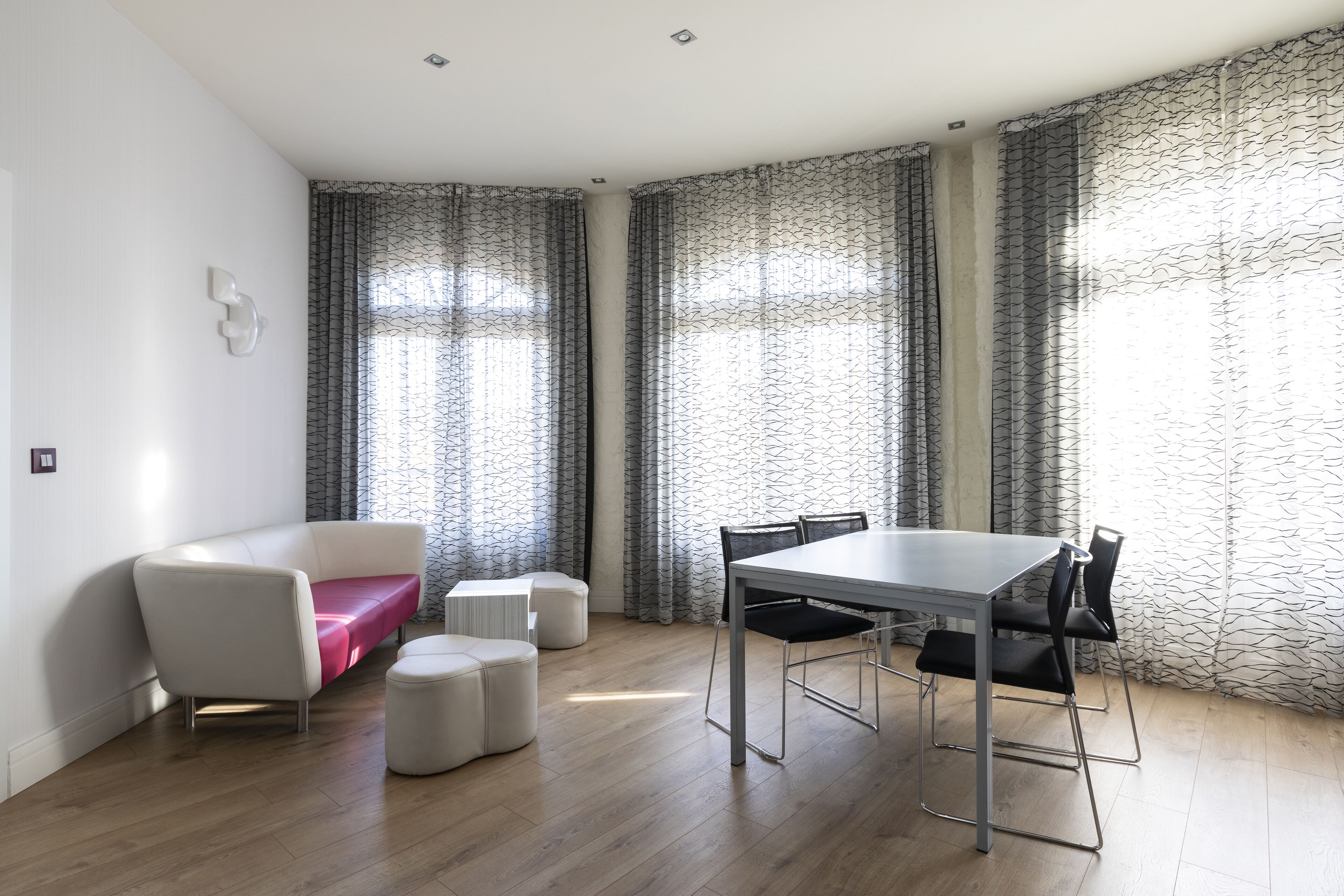 A modern living room with a white sofa, a dining table with chairs, and a patterned curtain.