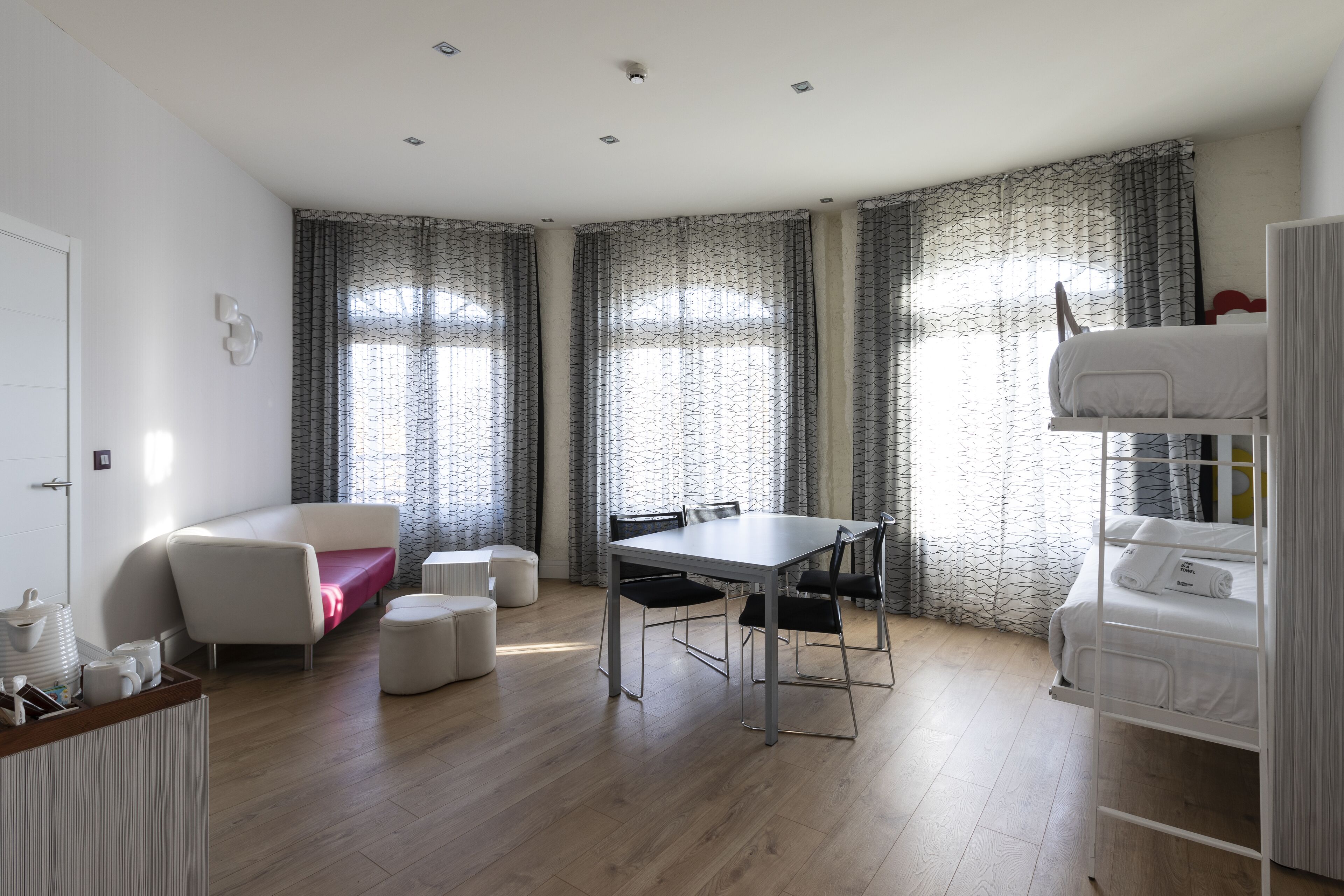 Una habitación moderna y luminosa con una cama blanca, una cama literera, una mesa de comedor con sillas, un sofá y una mesita con tazas y una bandeja.