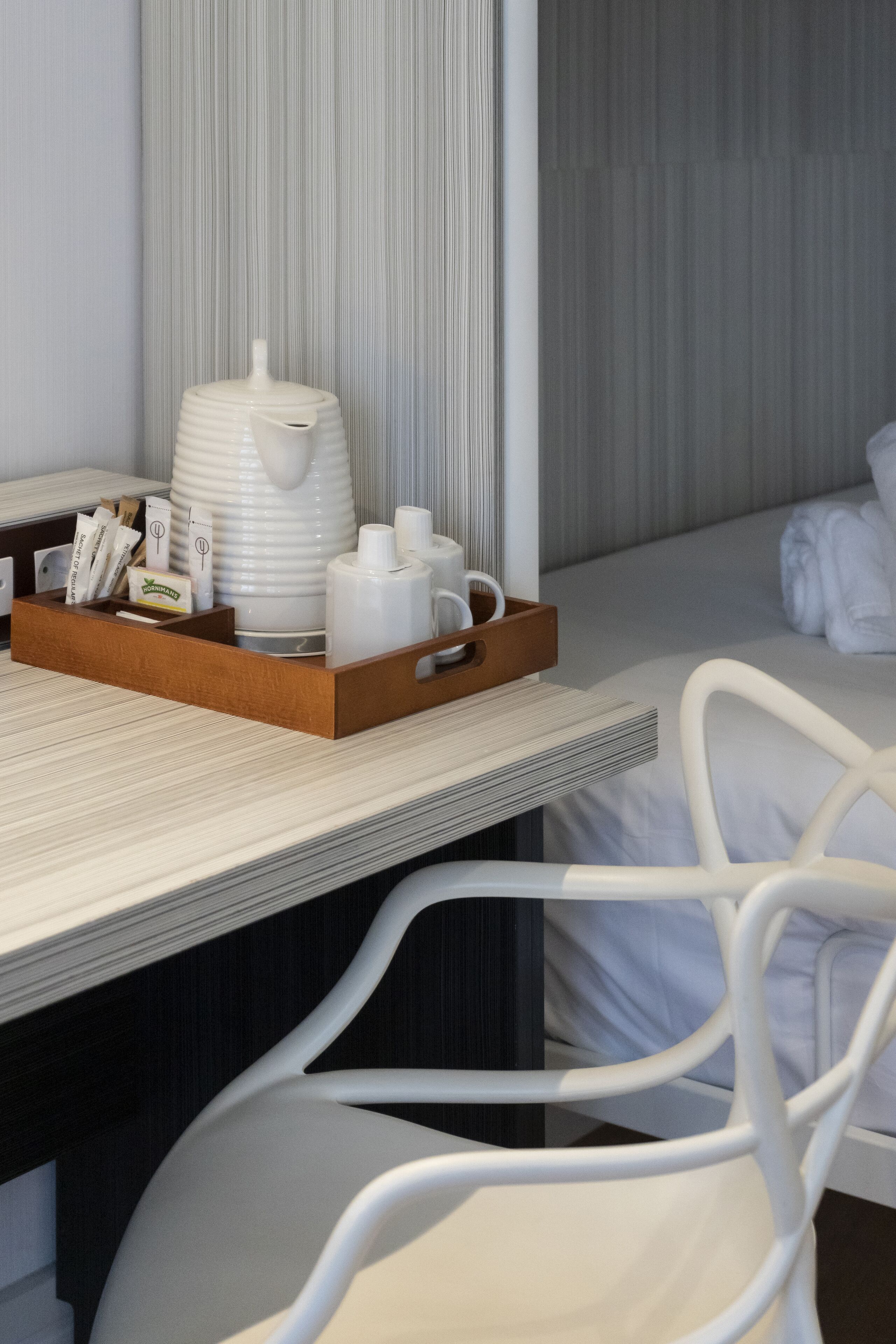 Habitación de hotel con una mesita de noche de madera que sostiene una tetera blanca, tazas y una bandeja con artículos de aseo. Una silla blanca se encuentra frente a la mesita, y se ve una cama con ropa blanca de cama al fondo.